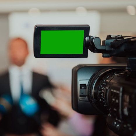 Video camera with a green screen, pointed at a blurred group of people indoors.