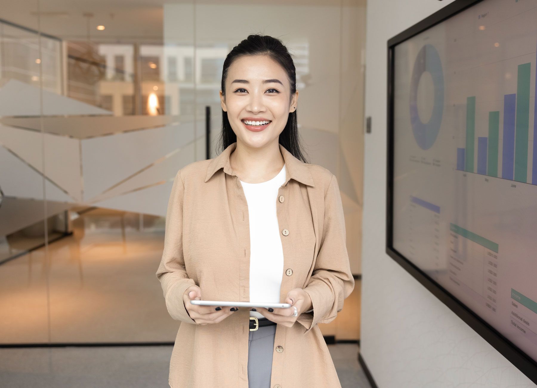 Woman holding a tablet smiles in front of a digital graph display, wearing a tan shirt and gray pants.