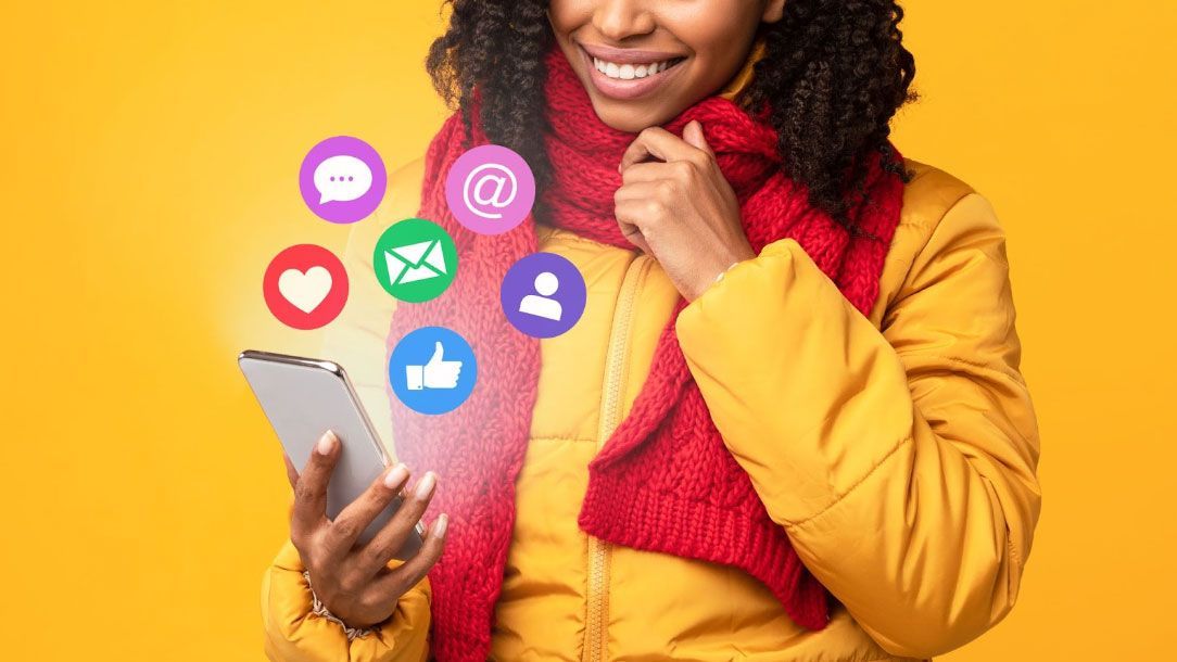 Smiling person in yellow jacket and red scarf, holding phone with social media icons emitting from it. Yellow background.