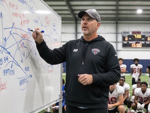 Football coach drawing plays on a whiteboard, explaining strategy to players. Indoor field setting.