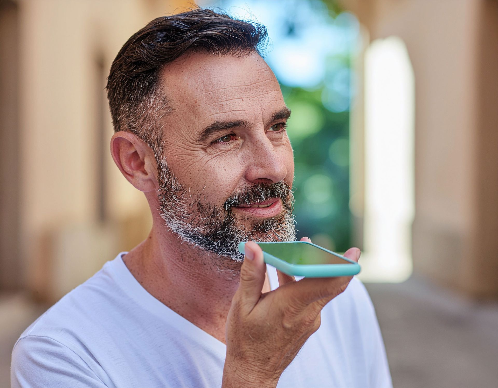 Man speaking into a phone, wearing a white shirt, outdoors with a blurred background.
