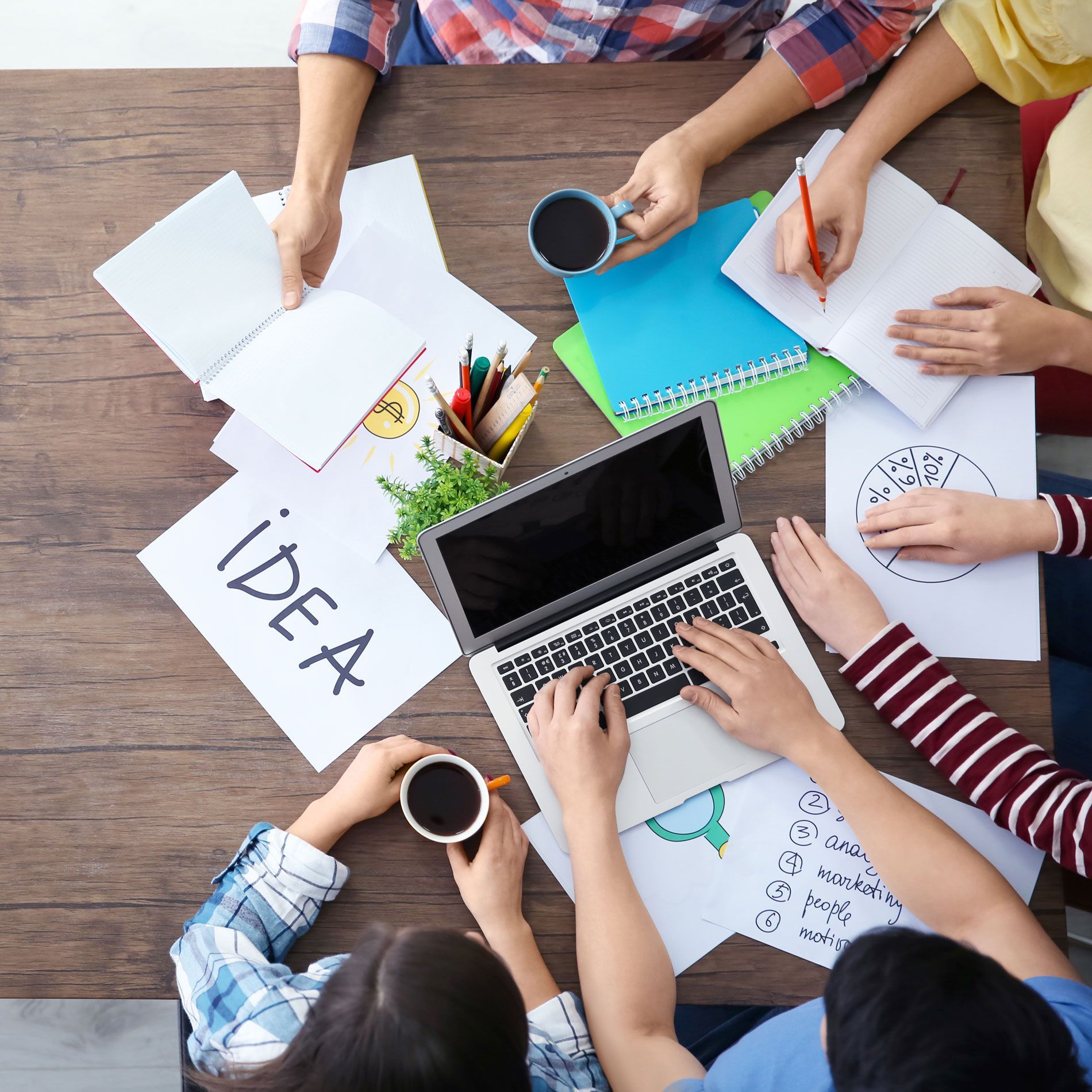 Group of people collaborating around a table, brainstorming ideas with notebooks, a laptop, and coffee.