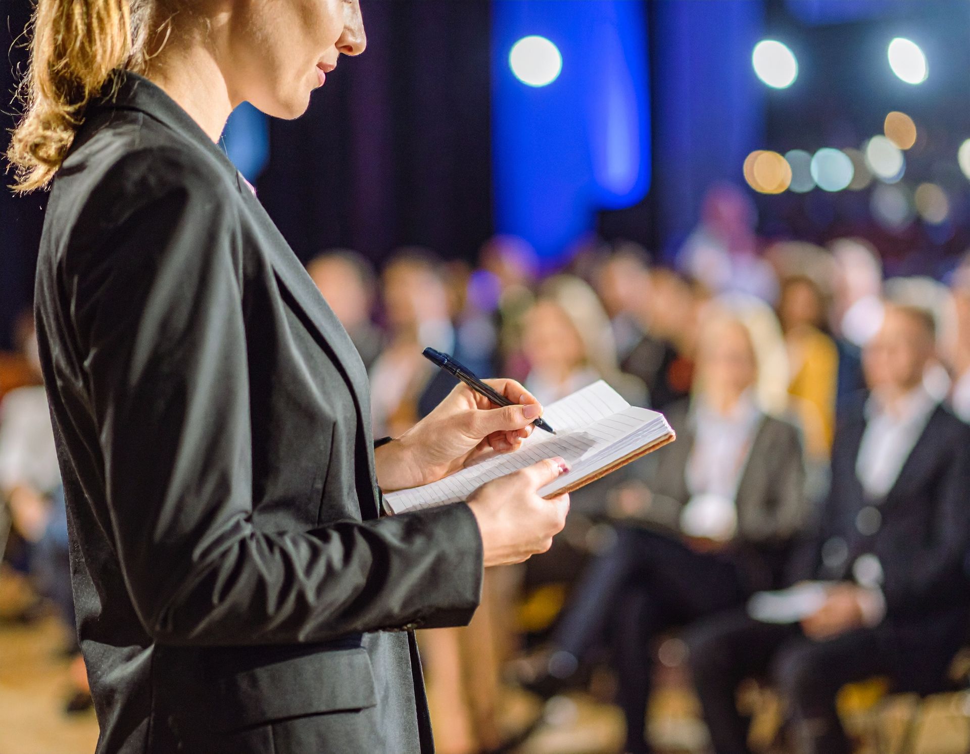 Woman in a black suit takes notes in a notebook during a conference; audience in background.