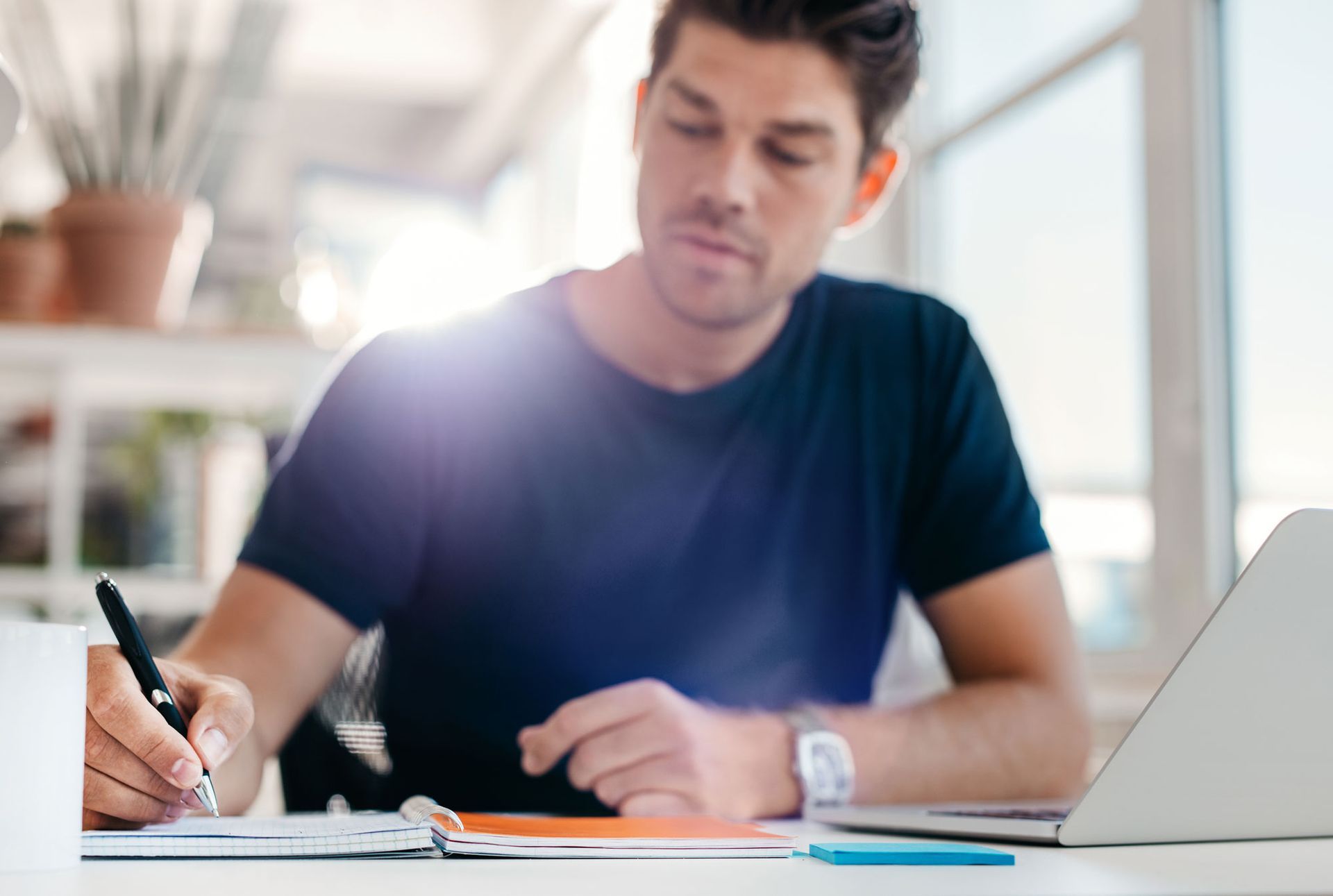Man writing in notebook with pen, laptop on desk, bright sunlight.