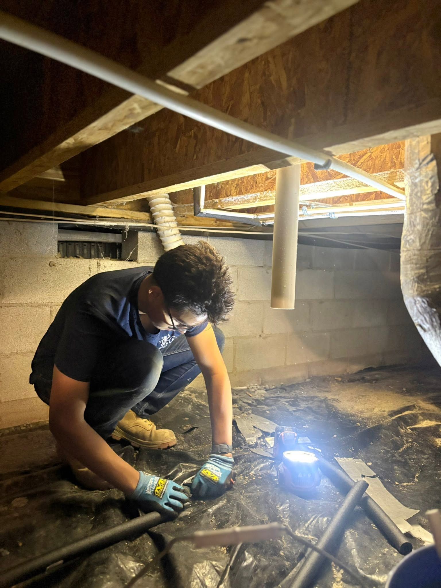 Person welding under a house in a crawlspace, wearing gloves and a face shield, with bright sparks lighting the area
