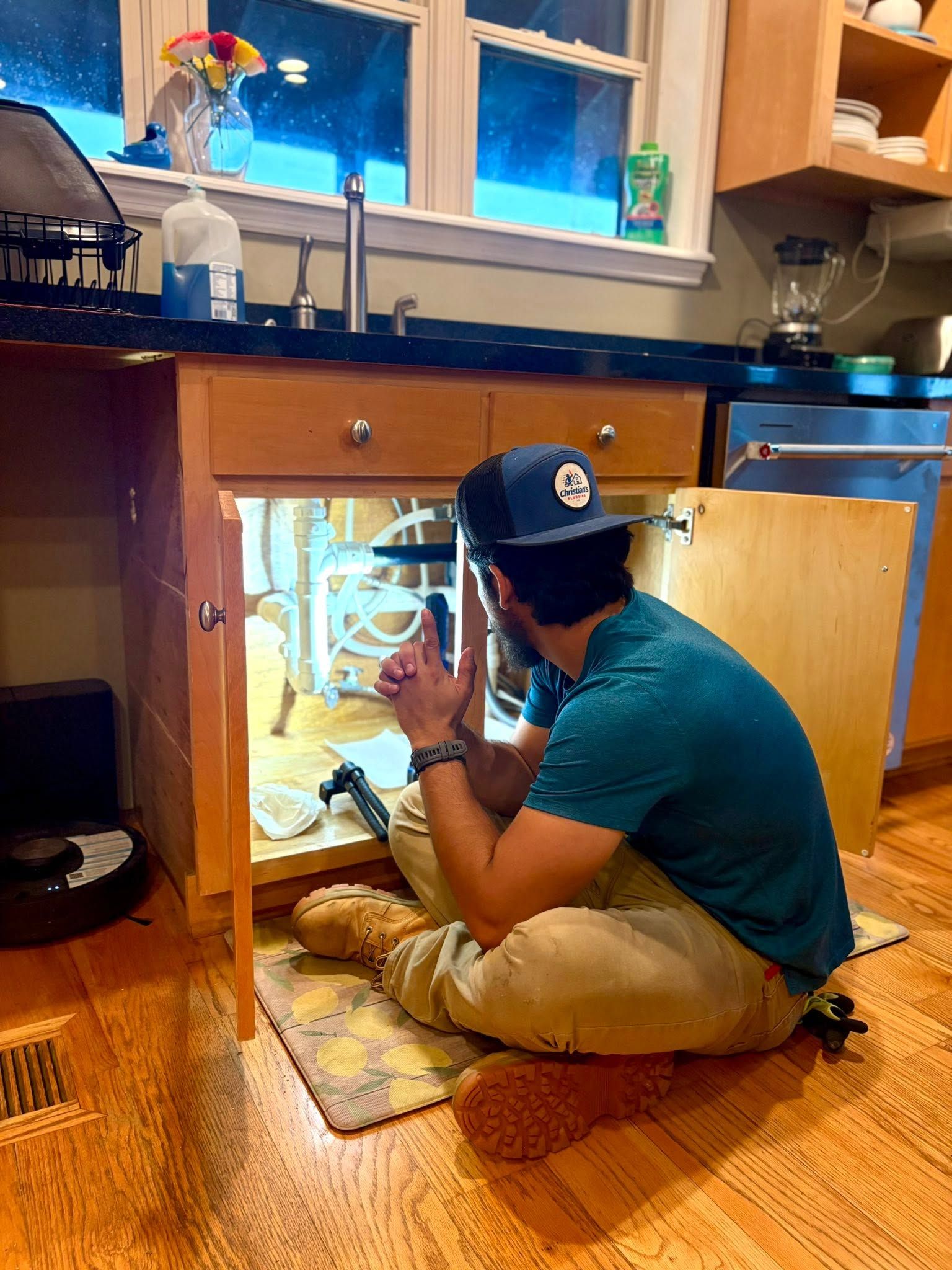 Person crouches under a kitchen sink, inspecting pipes with a flashlight.