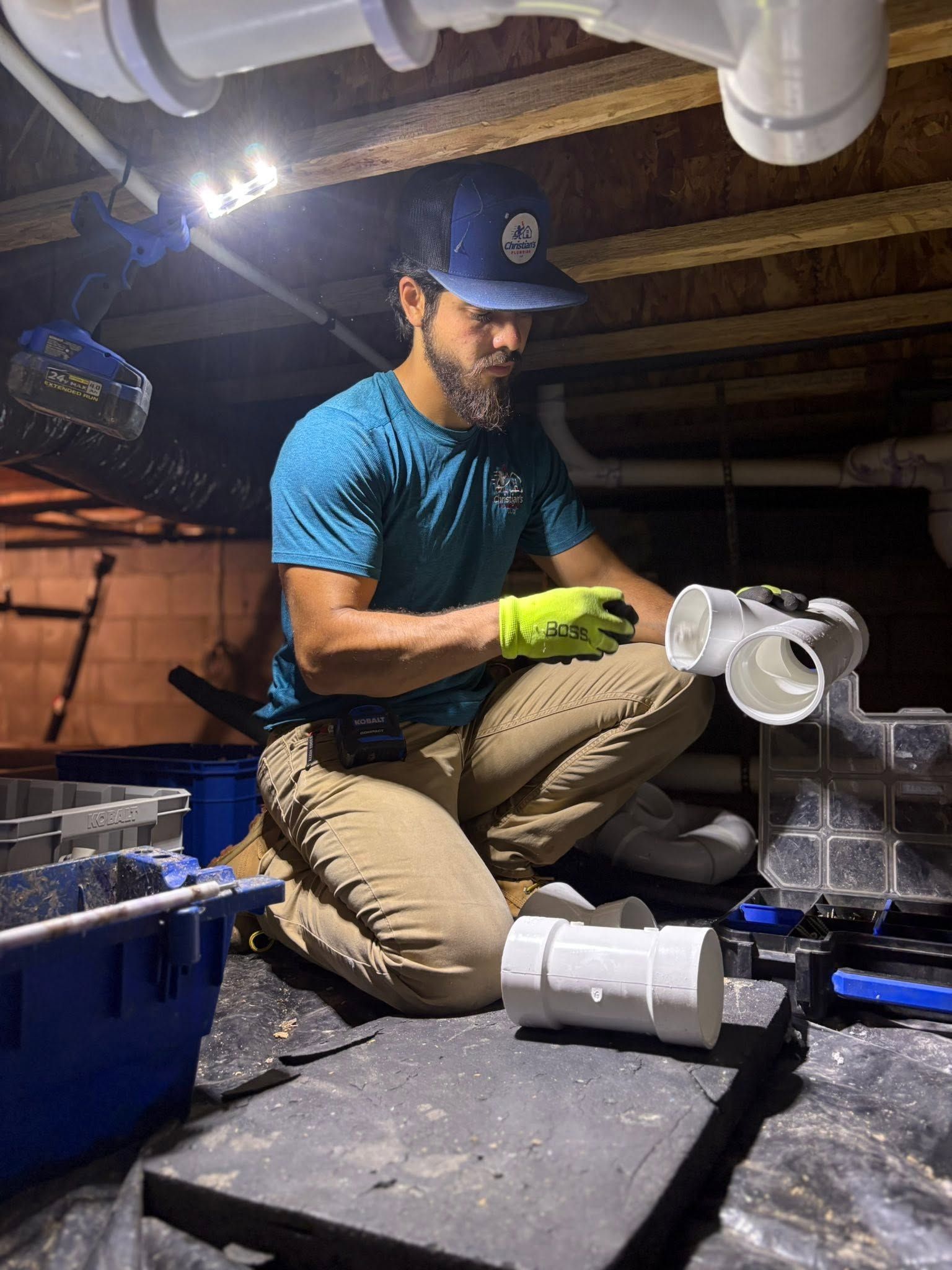 Person kneeling under a sink, installing white PVC pipes in a crawlspace while wearing gloves and a cap