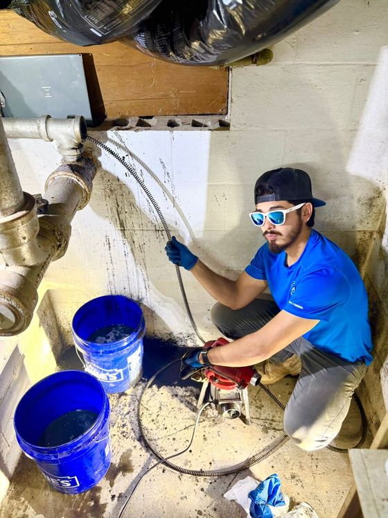 Worker in blue shirt repairs a ceiling pipe amid debris, with blue buckets and tools on the floor.