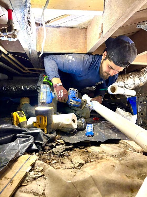 Worker crouched under floorboards, inspecting plumbing with tools in a cramped crawlspace.