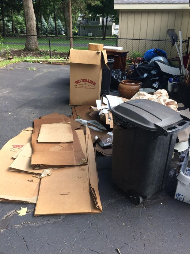 Pile of cardboard boxes, trash bin on a driveway, overflowing with discarded items, outdoors.