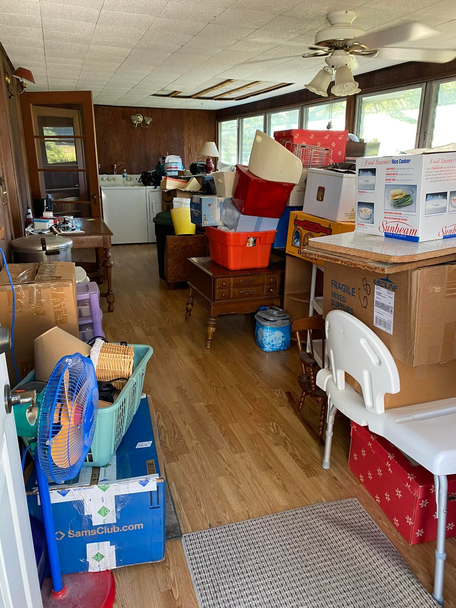 Cluttered room with boxes, furniture, and various items. Wooden floors and paneling; natural light enters.
