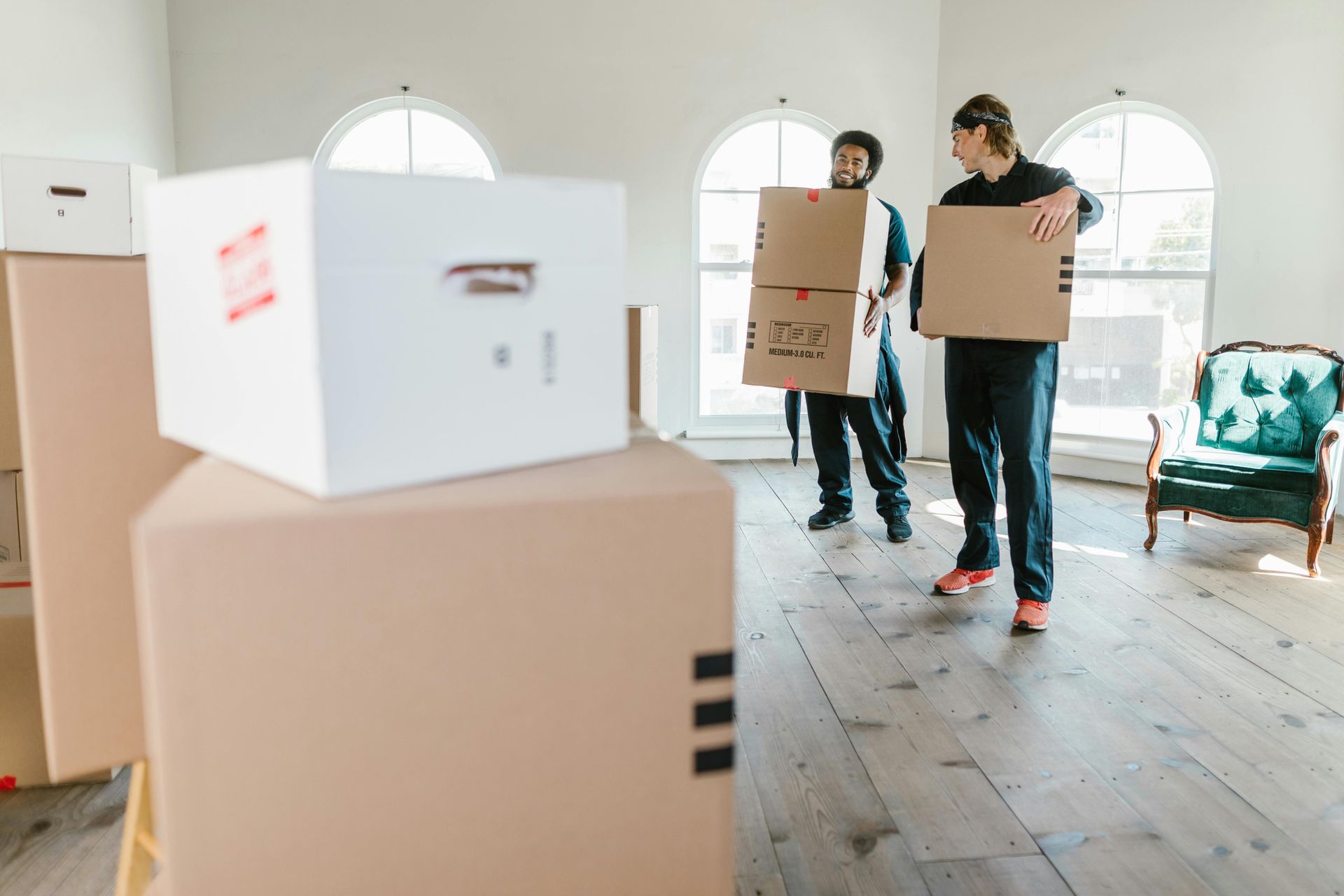 Two men carrying cardboard boxes into a room with a stack of boxes, a chair, and arched windows.