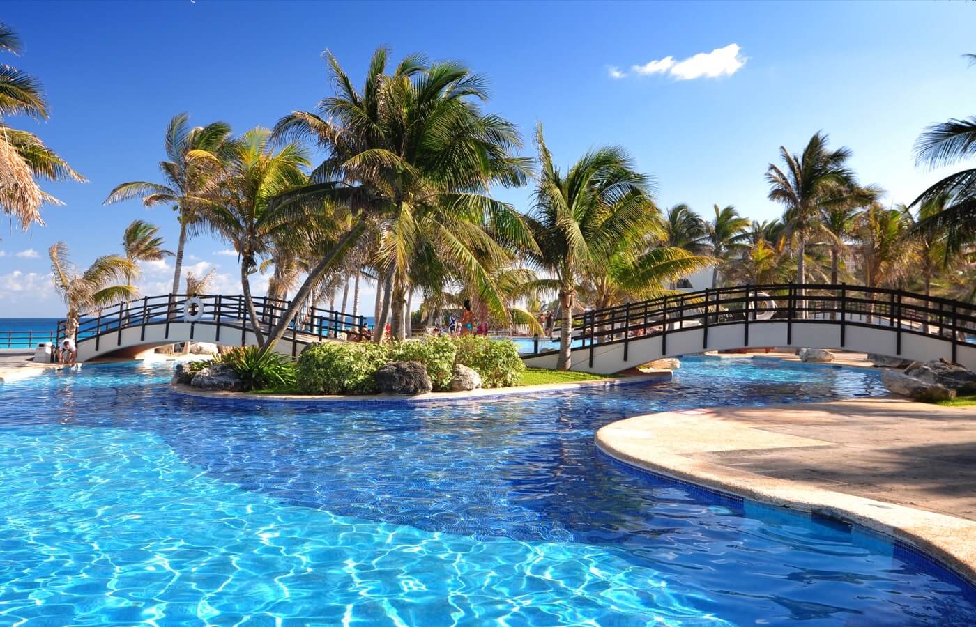 Hay un puente sobre una gran piscina rodeada de palmeras.