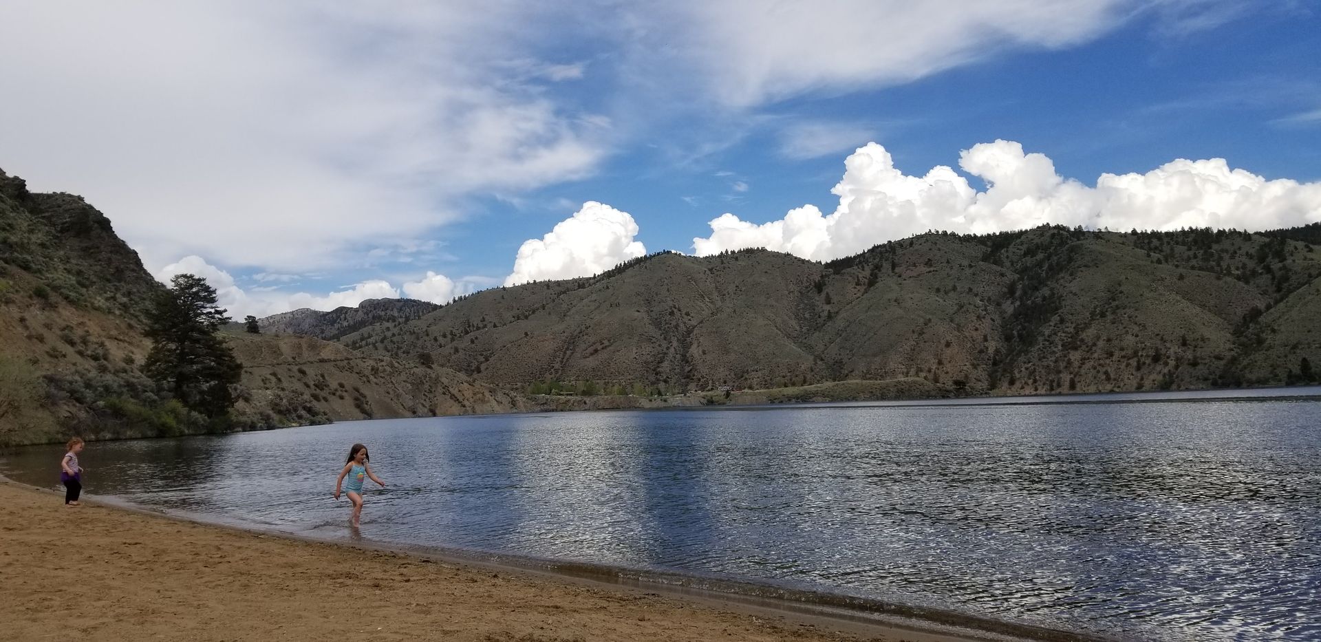 Lake with sandy shore, mountains, and blue sky with puffy white clouds. Two people stand near the water.