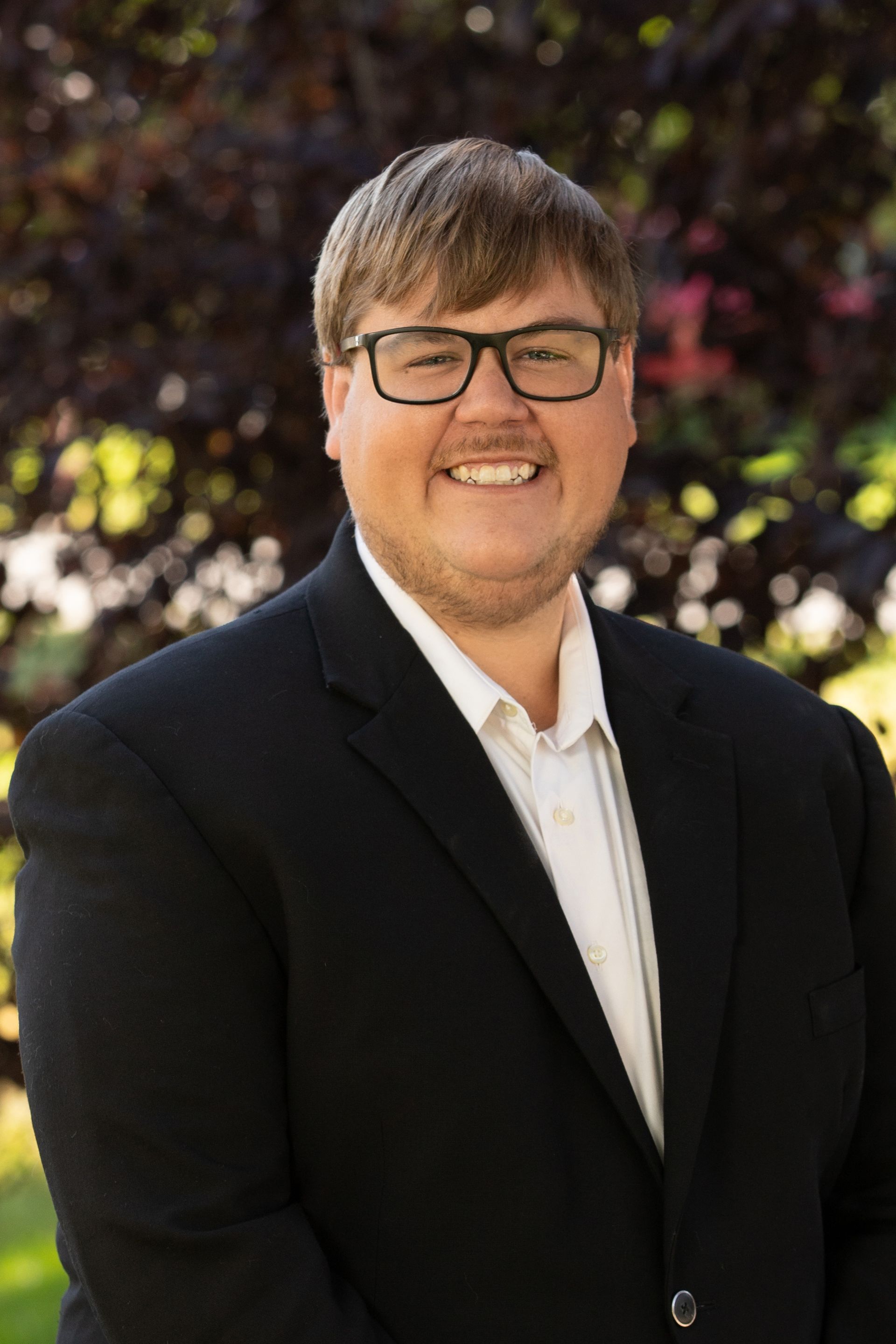 Woman with long, wavy hair, wearing a blazer over a white blouse, smiling outdoors.