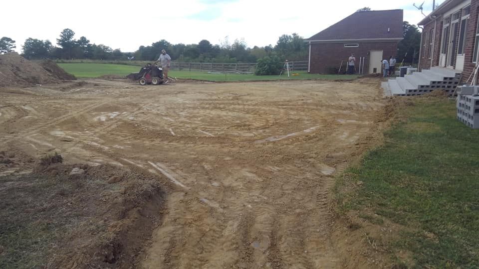 A tractor is driving through a dirt field in front of a brick building.