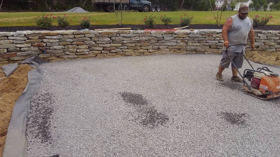 A man is working on a gravel driveway in front of a stone wall.