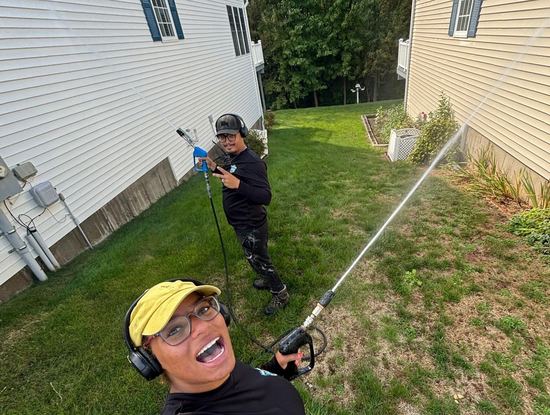 Two people power washing a house exterior; one poses smiling, the other points, both wearing safety gear, outdoors.