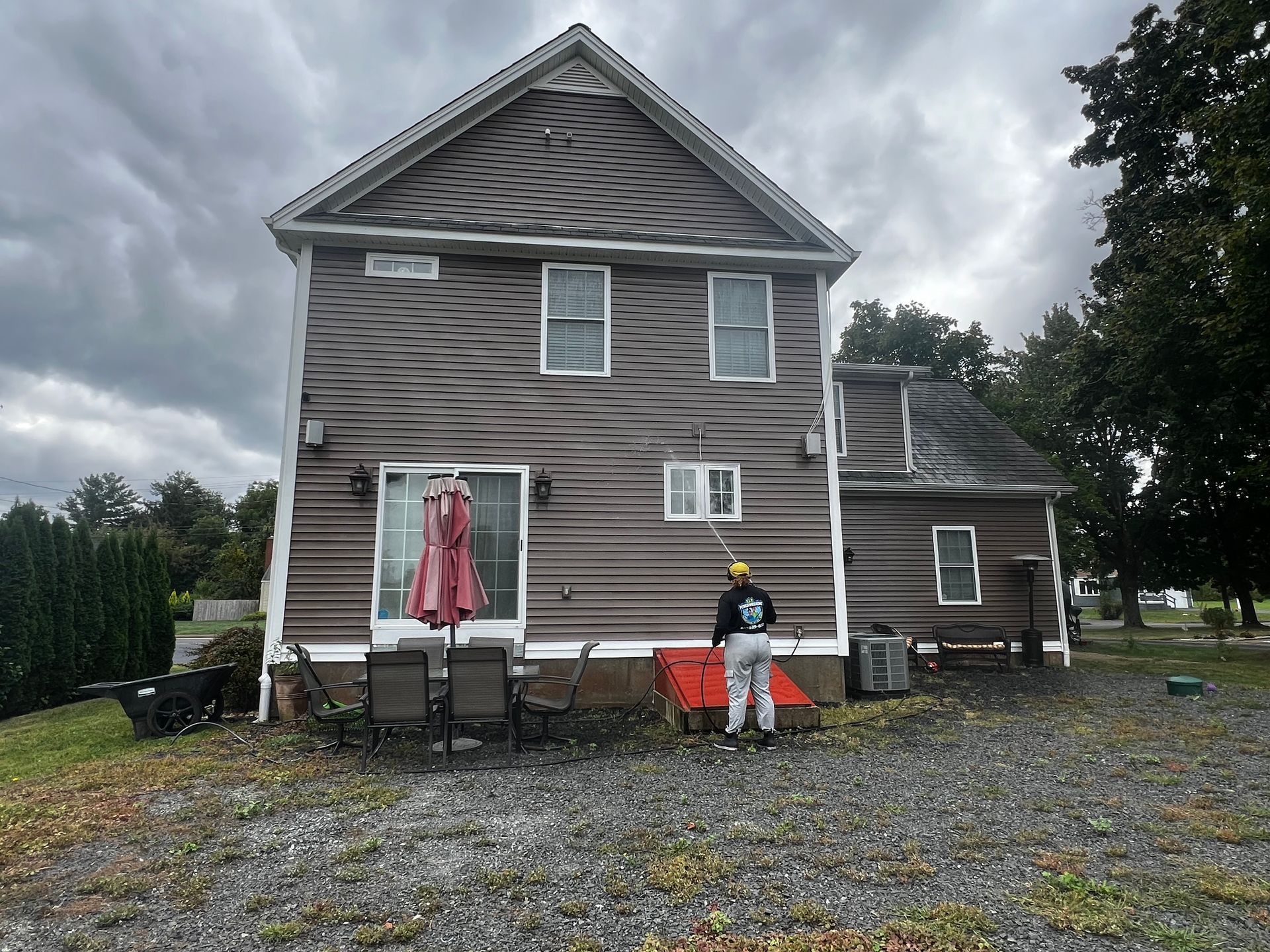 Brown house with a person painting the side. Cloudy sky, gravel yard, chairs, umbrella.