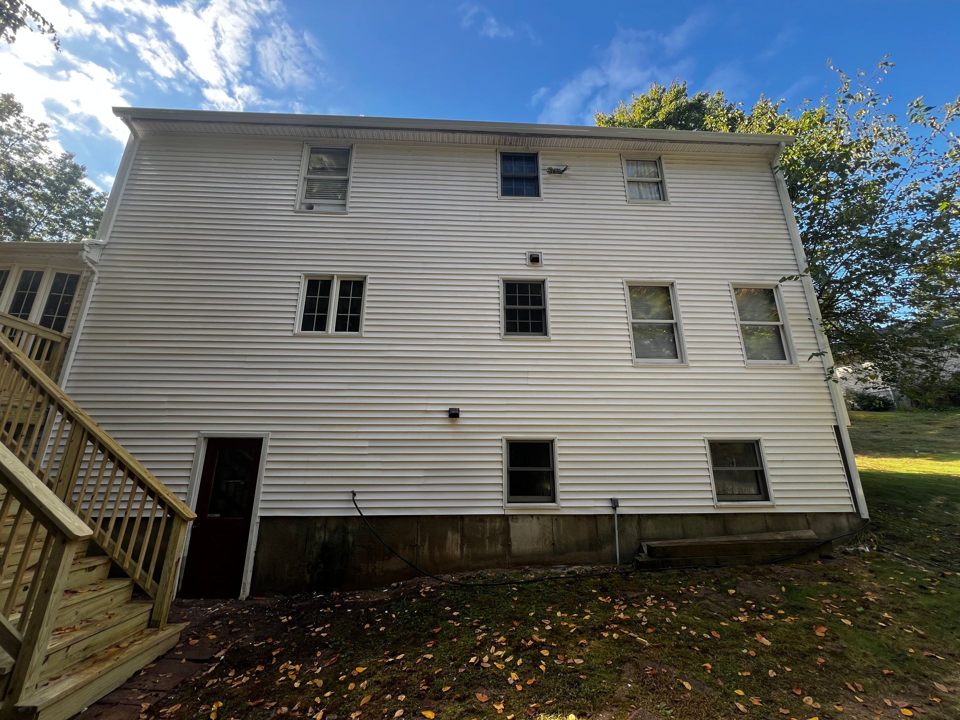 Back of a two-story white house with several windows. A wooden deck and yard with autumn leaves are visible.