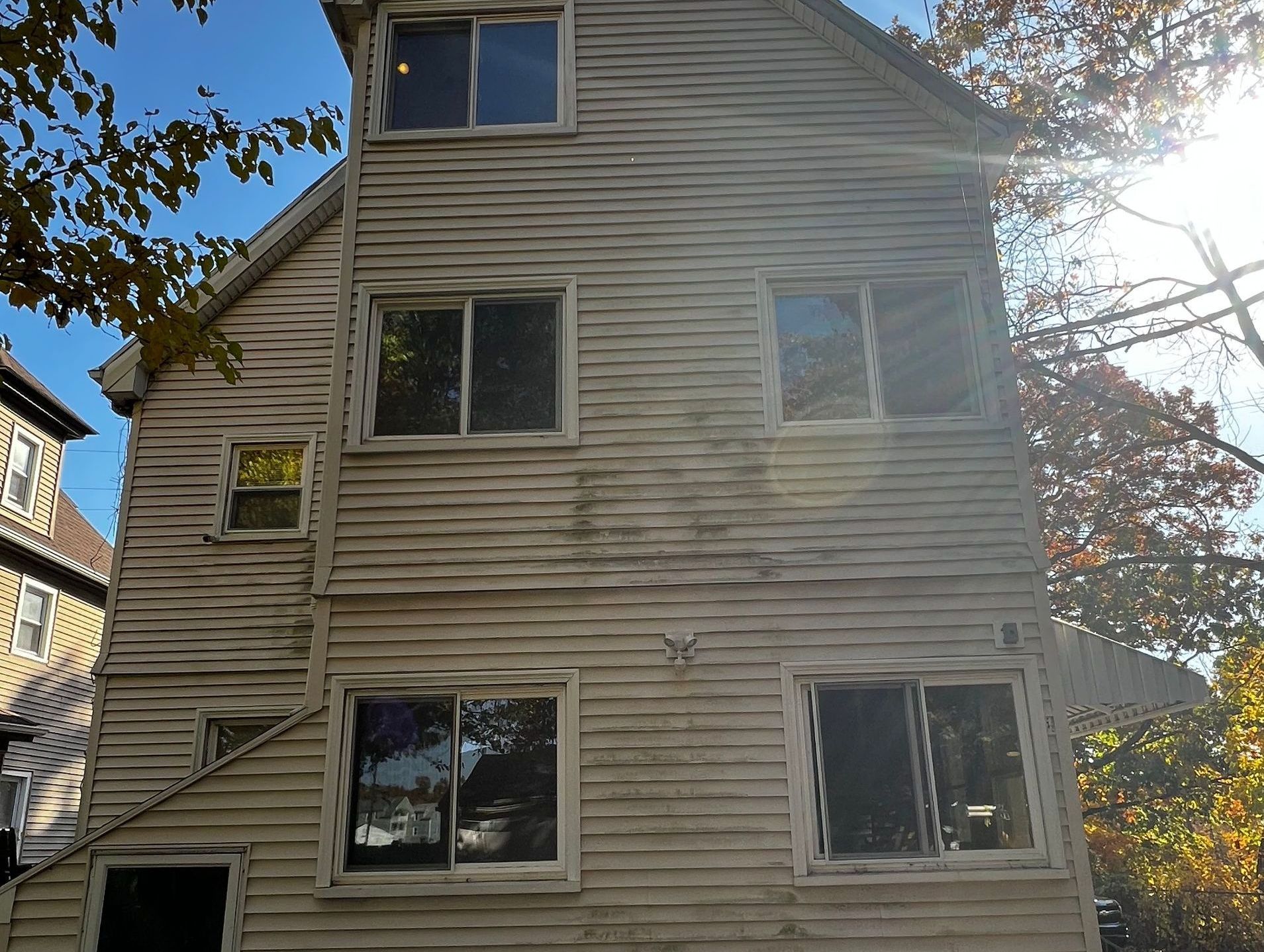Three-story weathered white building with multiple windows, branches and sun in background.