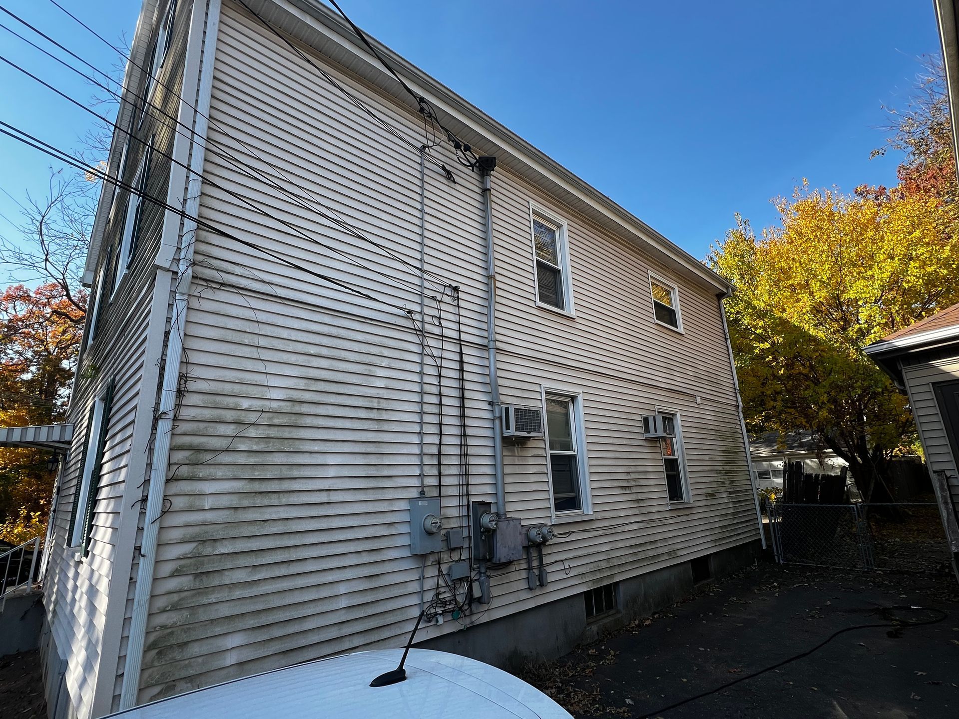 Side view of a two-story, light-colored house with electrical boxes and lines, blue sky, fall trees.