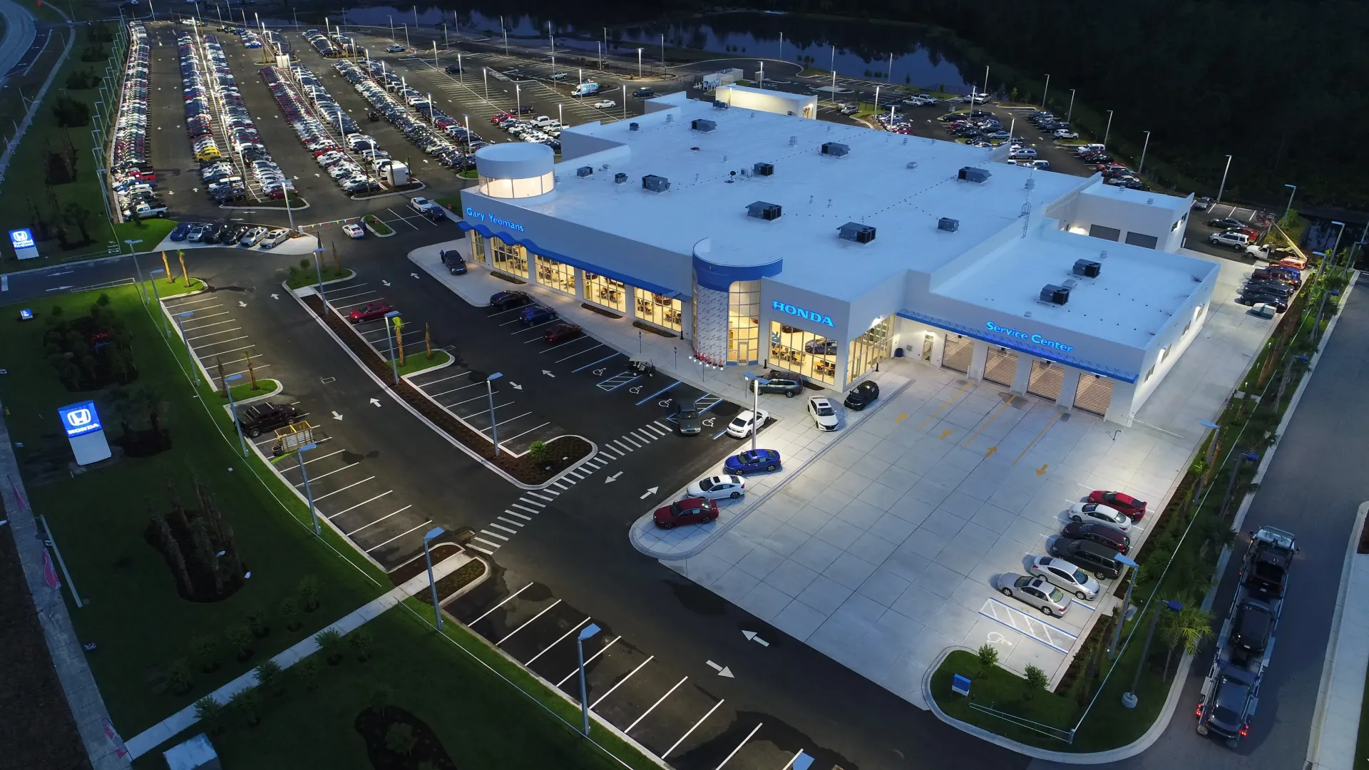 An elevated nighttime view of a brightly lit car dealership building with large parking lots filled with rows of vehicles.