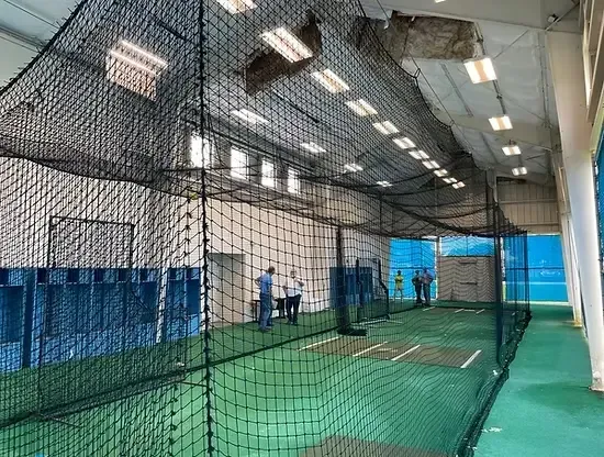 Indoor training facility with batting cages, green turf, netting, and three people standing inside.