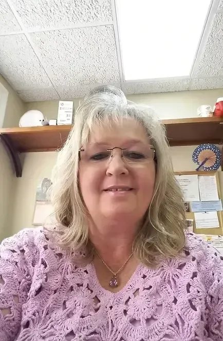Woman with blonde hair wearing glasses and a purple lace top, seated indoors.