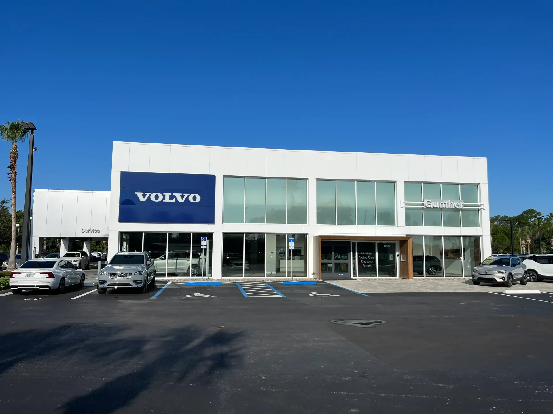 A bright white Volvo dealership building under a clear blue sky, with cars parked in the foreground lot.