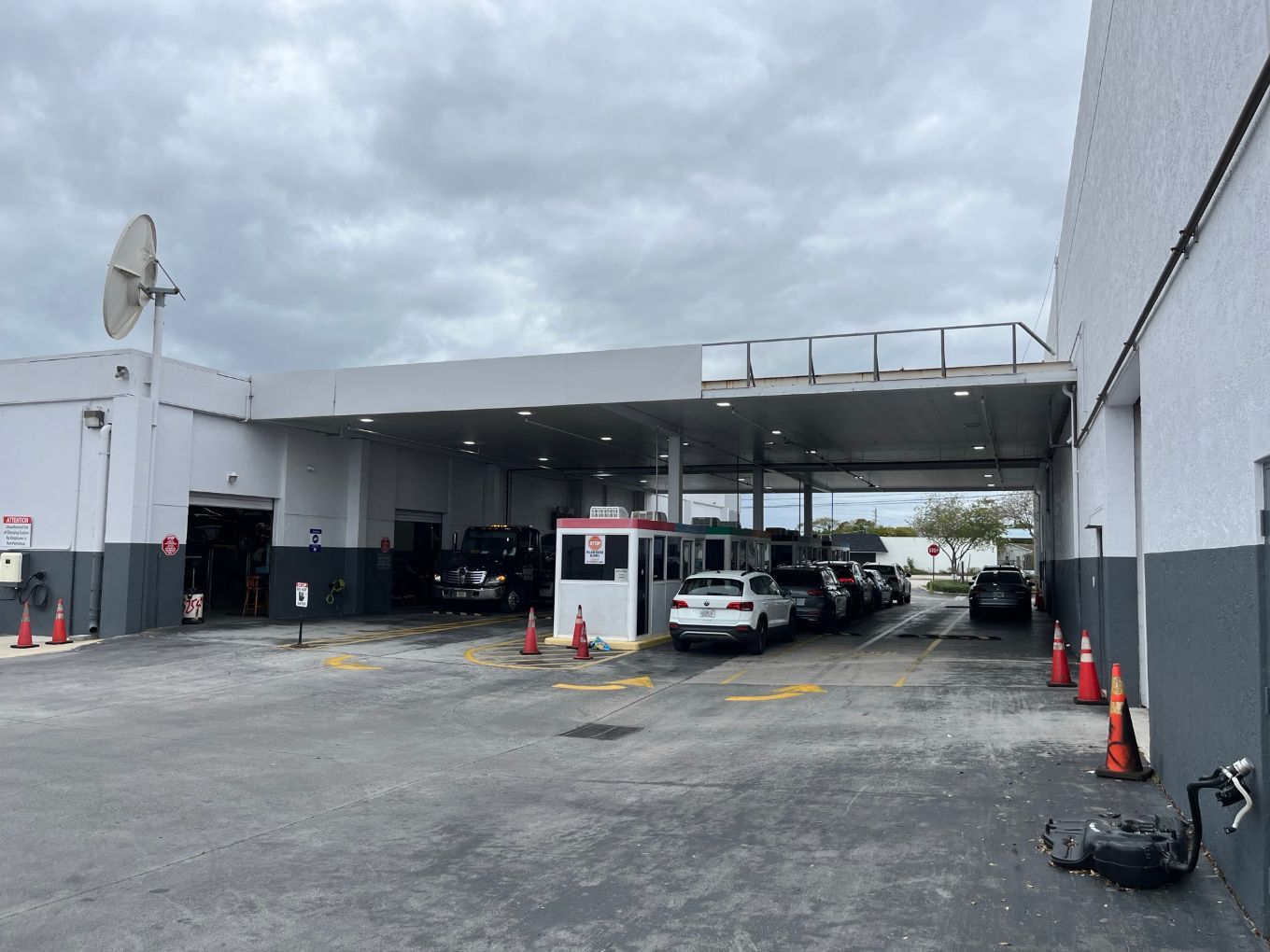 A white building with a covered service bay, parked cars, and traffic cones on a paved lot under a cloudy sky.