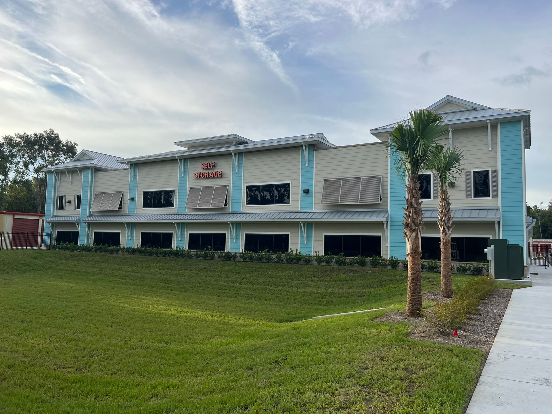 Two-story, light-colored building with blue accents, metal roof, and palm trees set against a green lawn and blue sky.