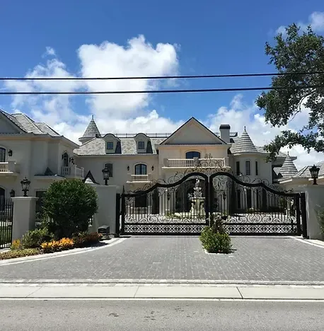 Ornate white mansion with a black gated entrance on a paved driveway, blue sky in background.