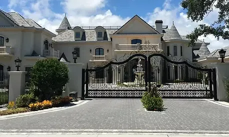 Large, ornate cream-colored mansion behind a decorative black iron gate with a paved driveway in front under a blue sky.