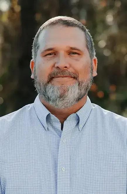 Man with salt and pepper beard smiles, wearing a blue patterned button-down shirt. Outdoor setting.