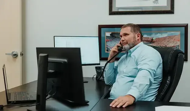 Man in blue shirt on phone in office, looking at computer screen, sitting at desk.