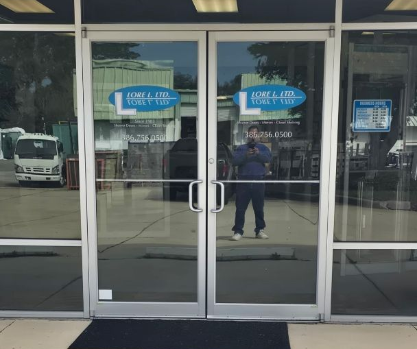 A set of glass double doors with blue signage at the entrance to a building, seen from a concrete sidewalk.