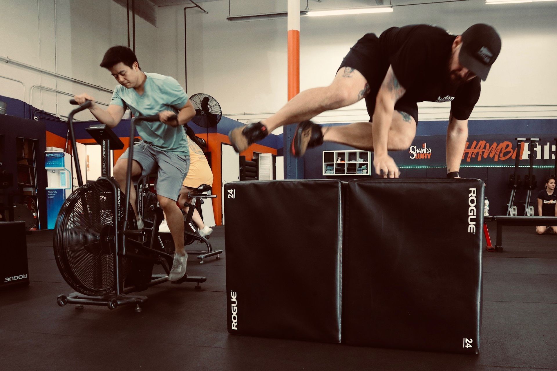 A man is doing a handstand while another man is riding an exercise bike in a gym.