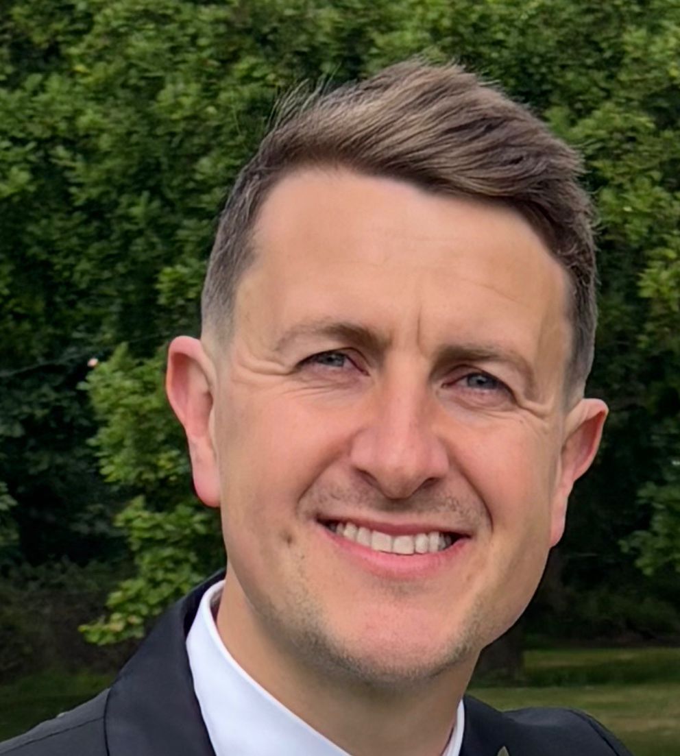 Man in a dark suit smiles broadly, with a backdrop of green trees.