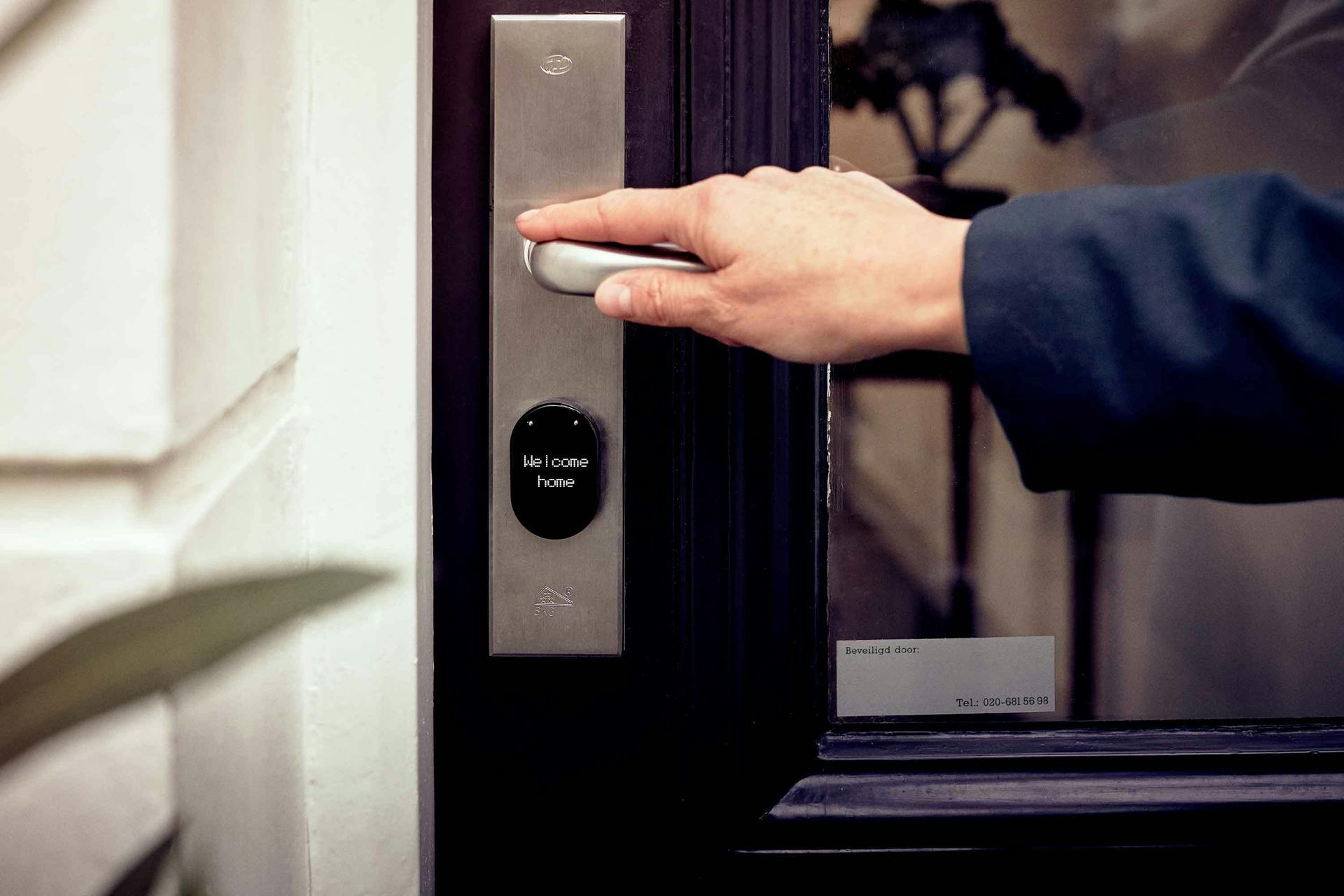 Person's hand on a door handle, opening a dark door with a key pad, next to a white wall.