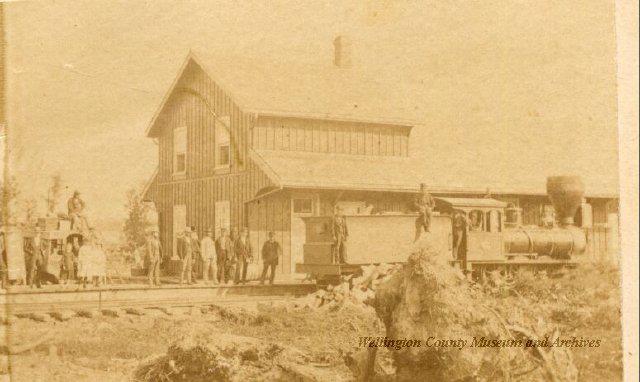 A black and white photo of a train station with people standing in front of it.