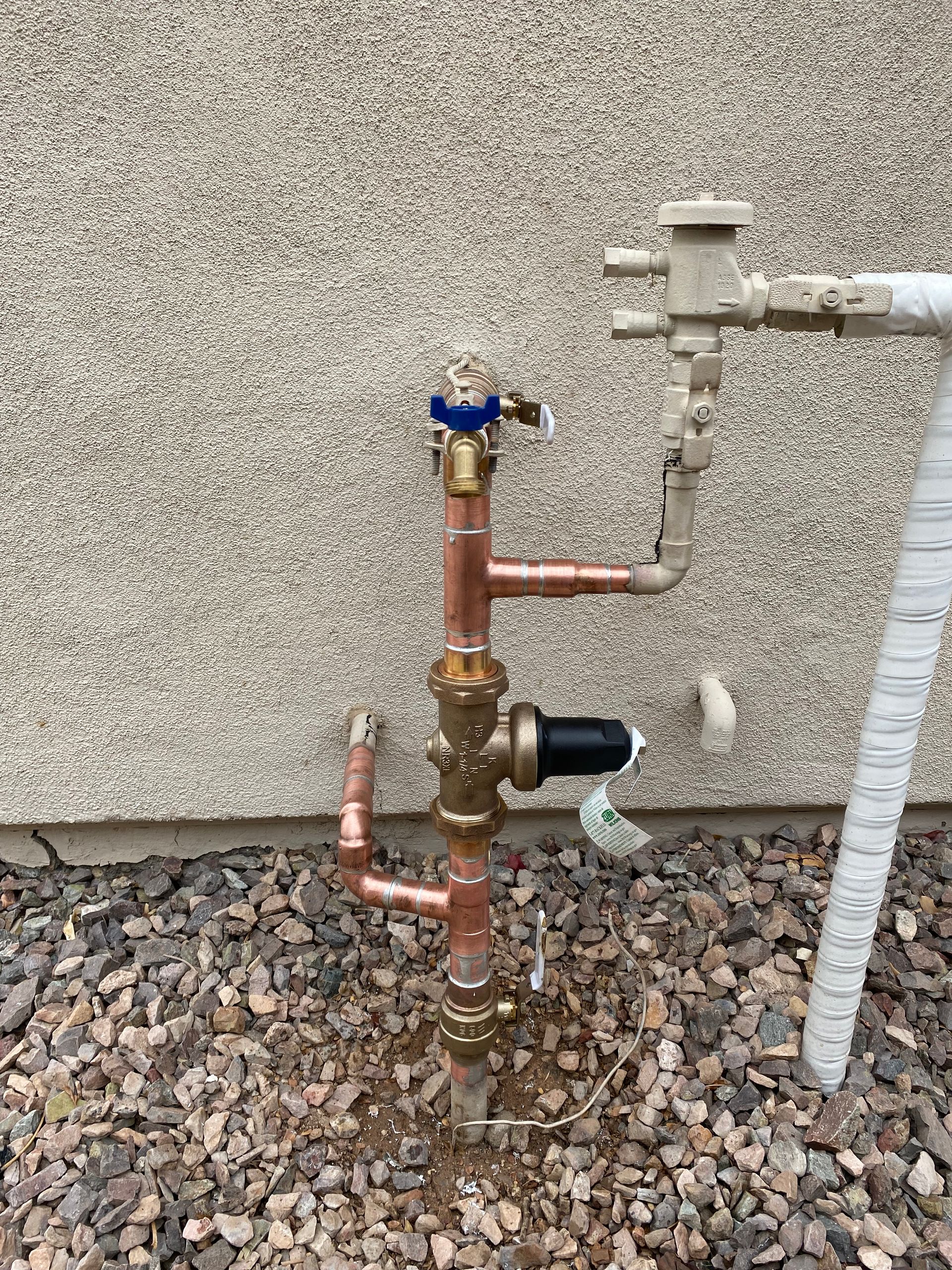 A copper pipe with a valve attached to it is sitting on gravel in front of a wall.