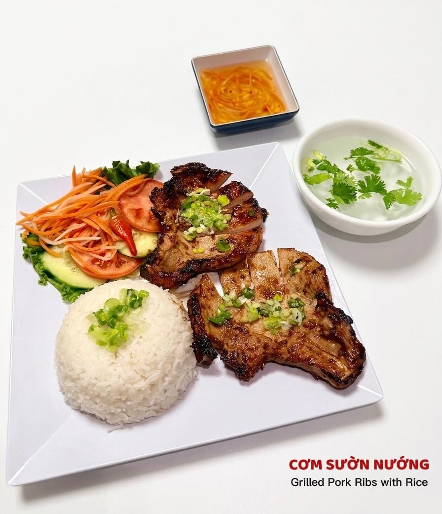 A plate of Vietnamese grilled pork chops with white rice, pickled carrots, cucumber, tomato, dipping sauce, and soup.