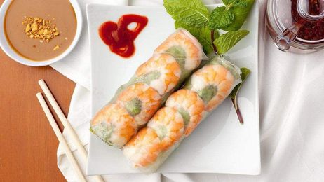 Two shrimp spring rolls with fresh mint on a white plate, served with a side of dipping sauce, chili oil, and chopsticks.