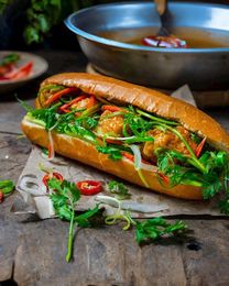A Vietnamese banh mi sandwich with meat, fresh cilantro, and chili slices on parchment paper, with a bowl in the background.