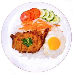 A plate of rice topped with a grilled pork chop, a fried egg, shredded carrots, tomato slices, and cucumber slices.