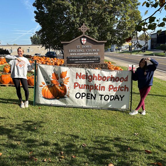 Two women at a pumpkin patch sign in front of a church. Fall setting with pumpkins.