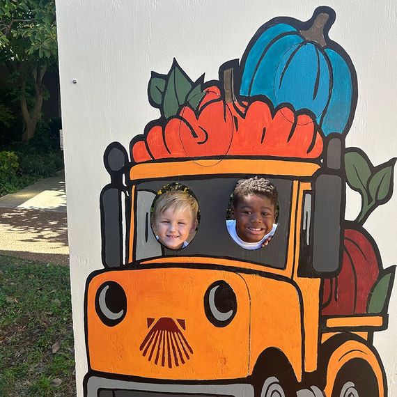 Two children with smiling faces, posed in a pumpkin truck cutout, orange and blue pumpkins above.