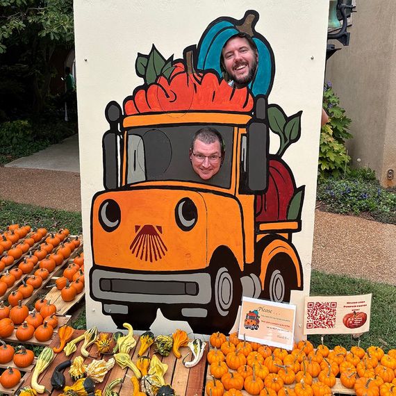 Two men pose with their faces in cutouts of an orange pumpkin truck. Pumpkins and gourds in foreground.