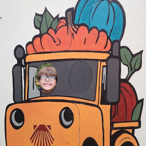 Child with glasses smiles, face in cutout on painted truck with pumpkins.
